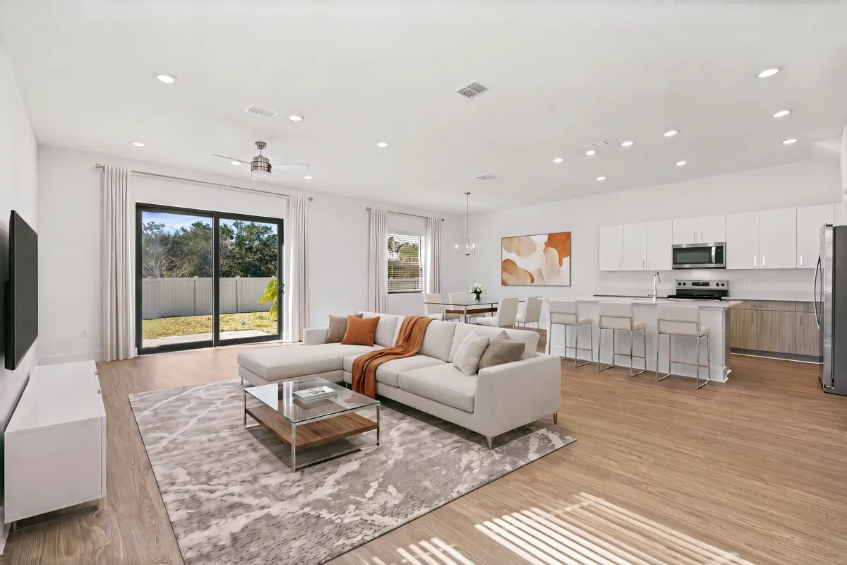 Maple great room with a sectional, a rug and a glass coffee table, the kitchen island and dining table behind it. Virtually staged.